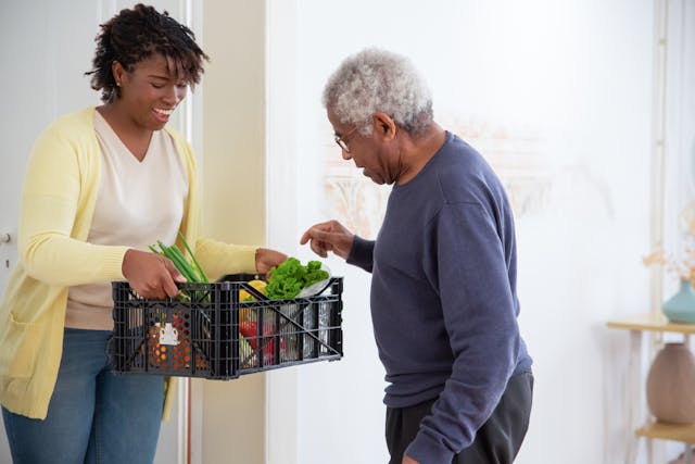A woman assisting an old man to pick fruit A young woman holding out a crate of vegetables to her grandfather